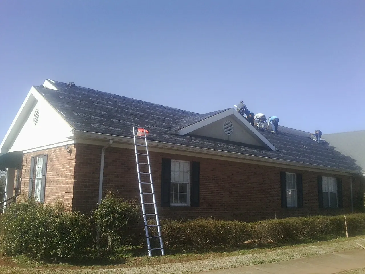 Crew tearing shingles off a brick ranch roof with a ladder set — Emergency Roof Repair work by our crew in Camillus
