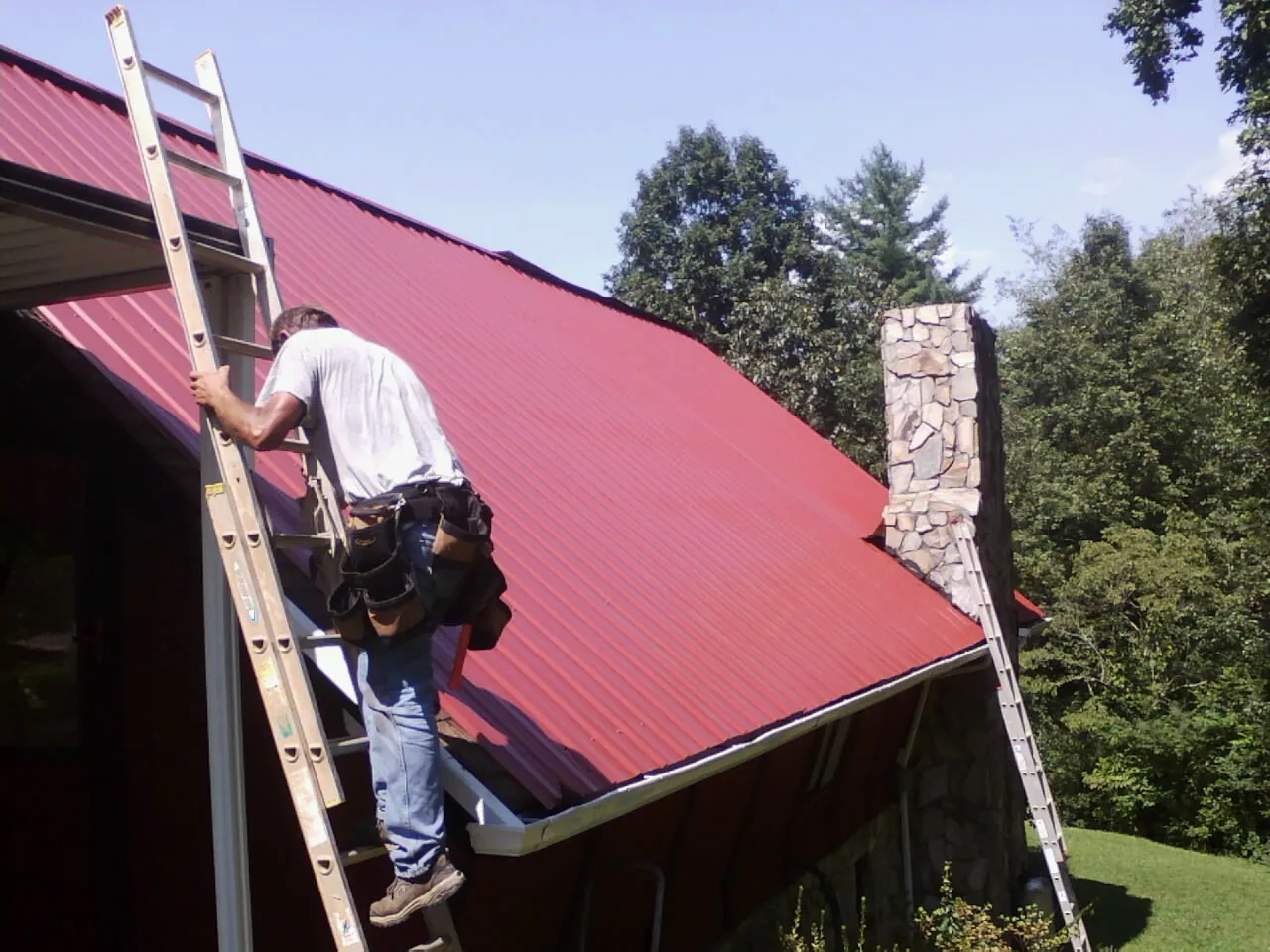 Roofer on a ladder beside a red metal roof and stone chimney — Soffit & Fascia Repair by Roofing Brothers in Camillus